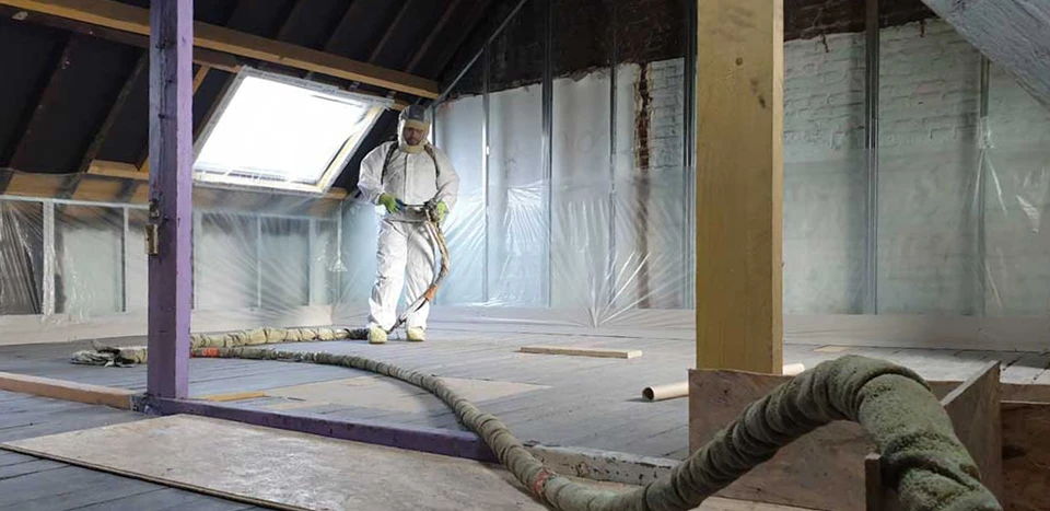 Technicien en combinaison blanche de protection intégrale applique de la mousse isolante polyuréthane projetée verte dans une zone de chantier confinée par des bâches plastiques fixées au ruban adhésif orange